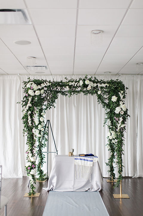 Wedding ceremony arch draped in white flowers and greenery against softly lit curtains, warm and romantic atmosphere