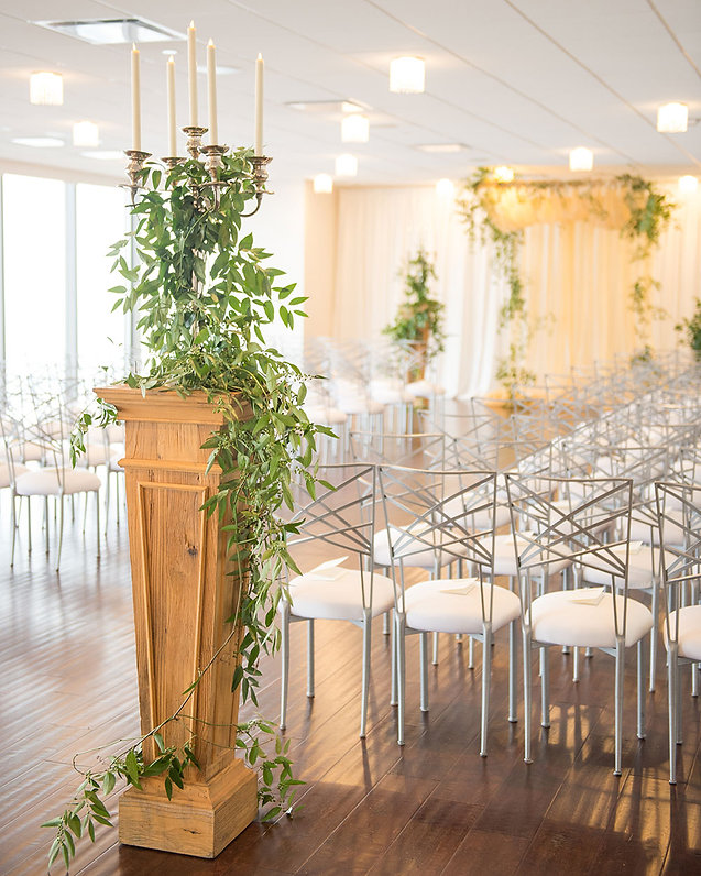 Analog film photograph of a wooden pedestal with cascading green vines and candles beside white chairs, bright airy venue
