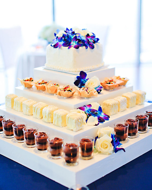 Tiered white dessert display with a small cake topped in blue orchids, white pastries, and chocolate dessert cups, soft creamy tones