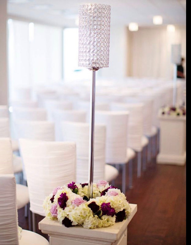 Tall crystal-topped candle stand decorated with white and purple flowers beside rows of white draped chairs in a bright banquet hall, analog film grain, warm ambient glow