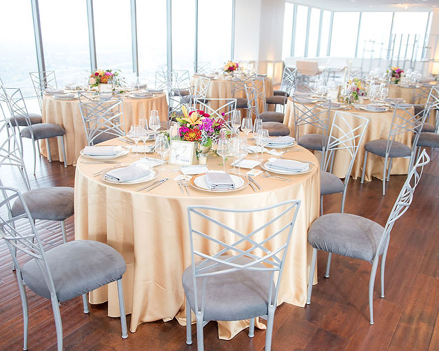Banquet room with round tables in champagne linens, gray chairs, floral centerpieces and floor-to-ceiling windows overlooking a city