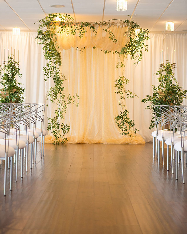 Indoor wedding ceremony setup with a draped gold fabric backdrop, greenery vines, candelabras, and rows of white chairs in a warm-lit venue, analog film grain